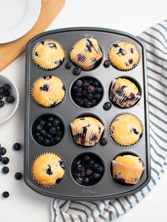 Several blueberry muffins in muffin tin; some muffins turned on their side and some cups filled with fresh blueberries.
