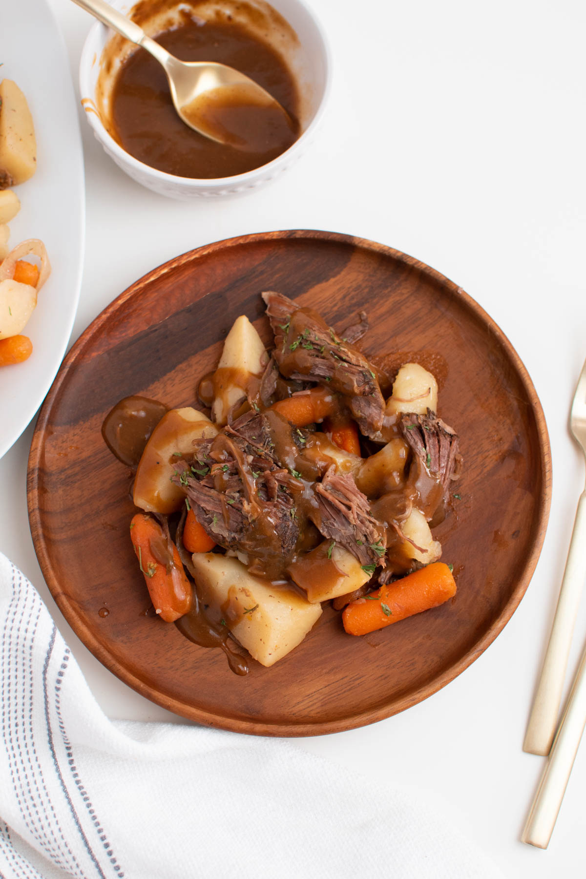 Pieces of pot roast, carrots, potatoes, and gravy on wood plate, which is on white table.