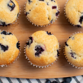Several blueberry muffins arranged neatly on wood cutting board with striped towel underneath.