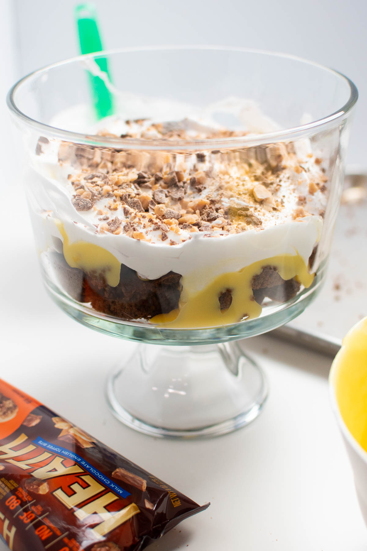 One layer of gingerbread trifle with whipped cream in glass serving bowl with bag of toffee bits nearby.