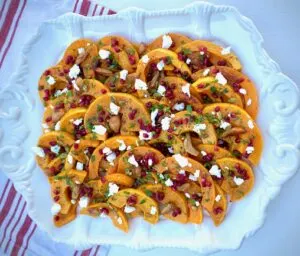 Roasted butternut squash with pomegranate and cheese with green garnish on white platter.