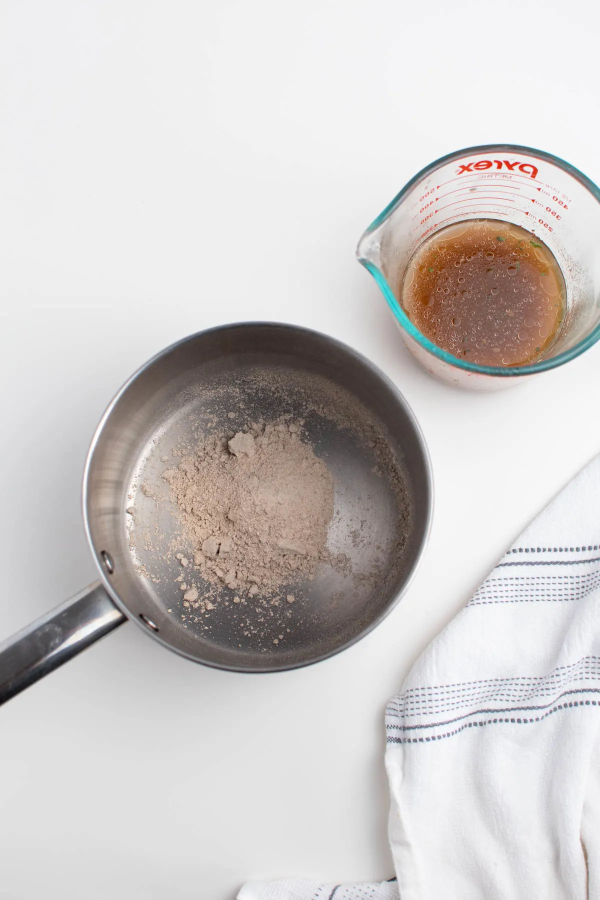 Small sauce pan with powdered gravy and glass measuring cup of beef broth on white table.