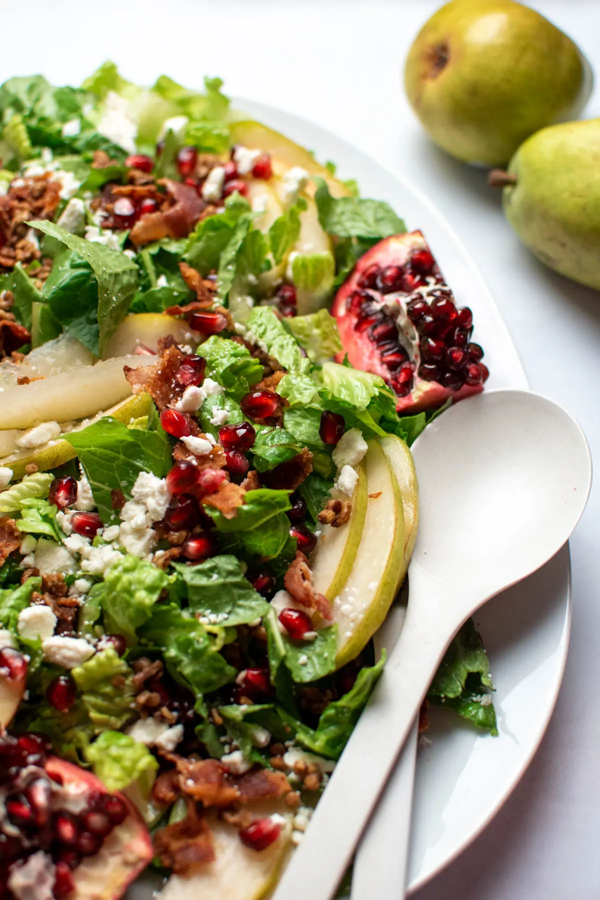 Green salad with pomegranate seeds and pears on white platter with serving utensils.