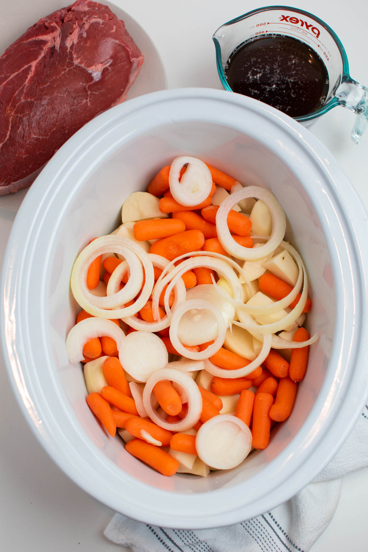Raw onion rings and baby carrots in white Crock Pot insert with other ingredients nearby.