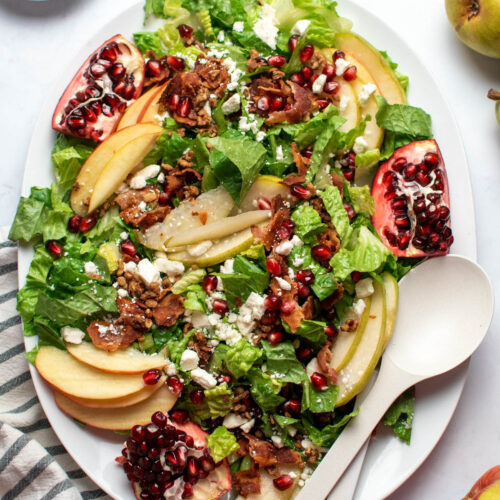 White platter with winter salad with pears, apples, pomegranates, and serving utensils on countertop.