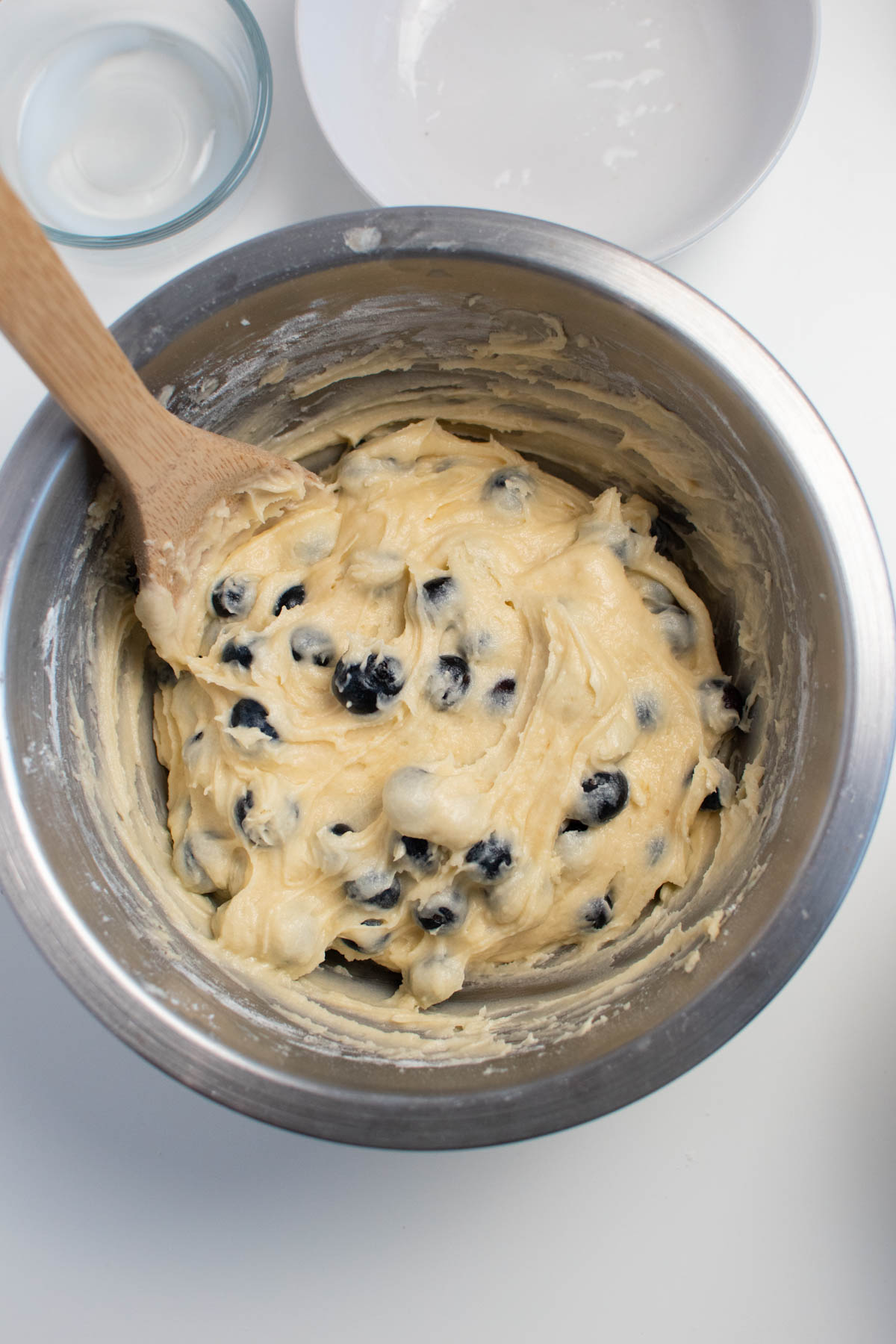 Blueberry muffin batter in metal mixing bowl with wood spoon in batter.