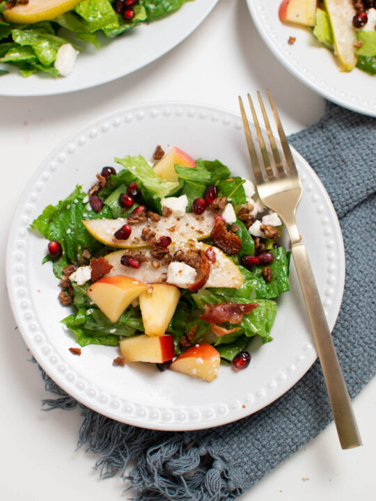 White plate with winter salad of lettuce, pears, pecans, and pomegranate seeds; gold fork resting on plate.