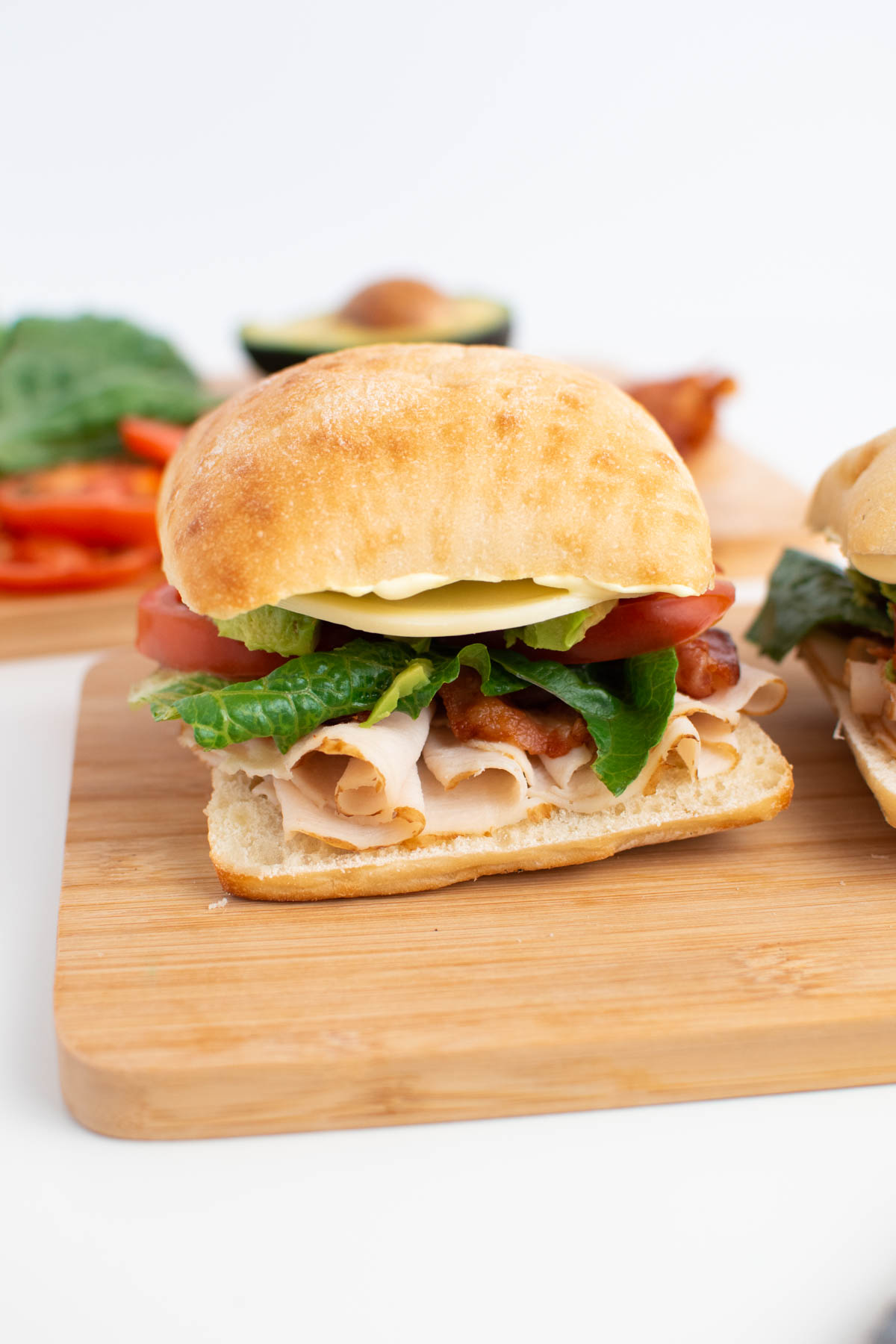 Avocado turkey sandwich on wood cutting board with sandwich ingredients in background.