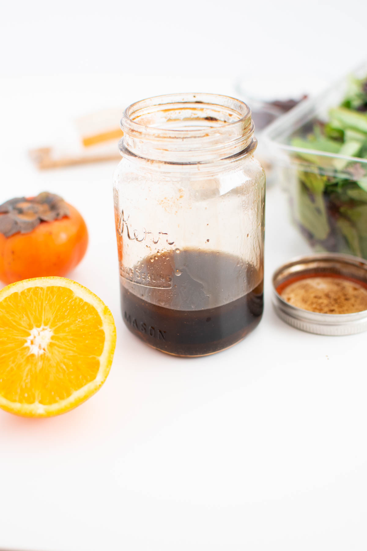 Glass jar with a small amount of balsamic dressing on white table with fresh persimmon and orange nearby.