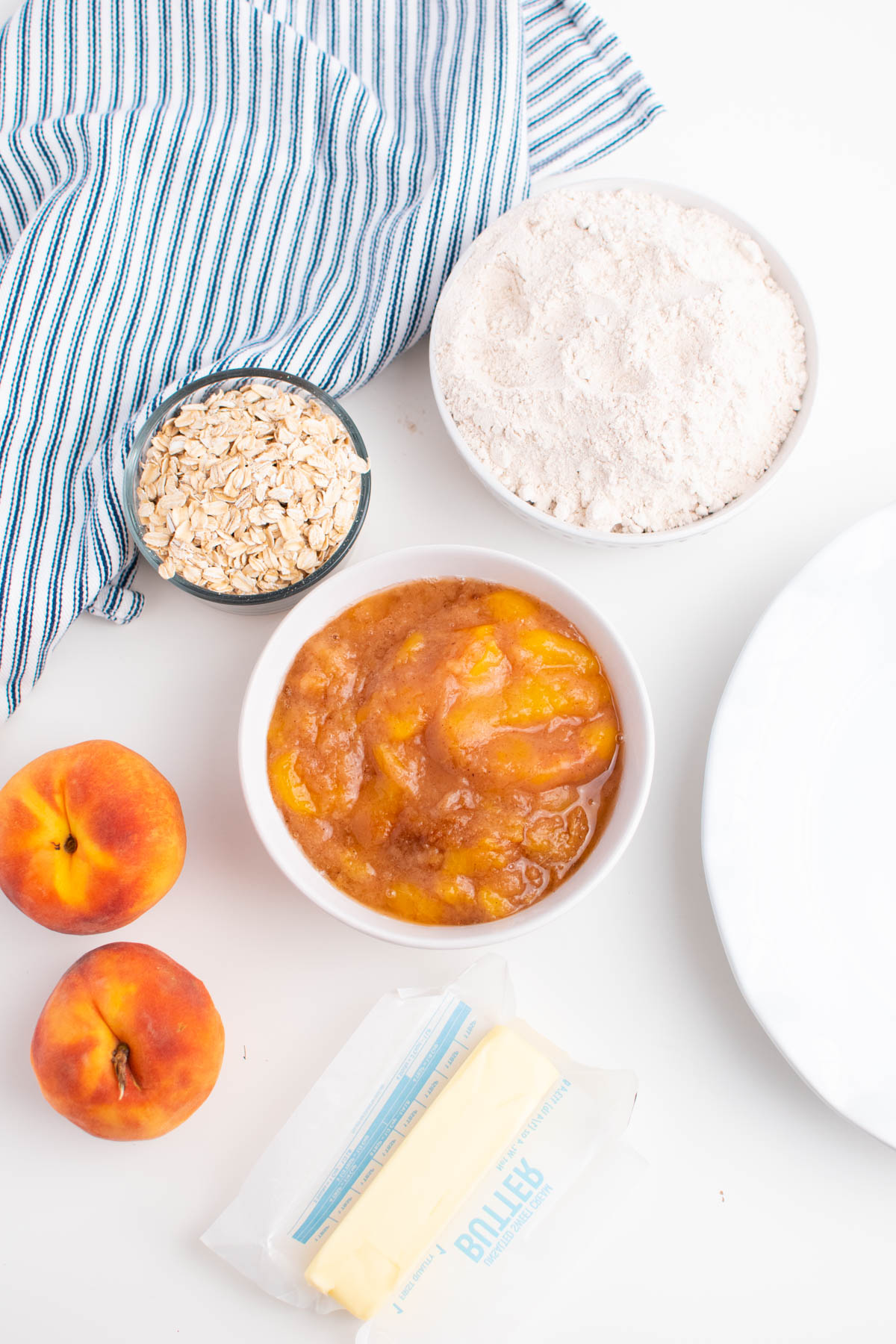 Bowls of peach crisp ingredients on white table including butter, cake mix, and peaches.