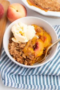 White bowl with peach crisp and scoop of vanilla ice cream with blue striped towel underneath bowl.