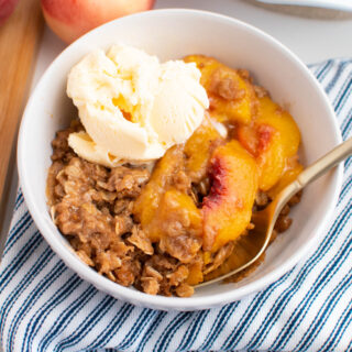 White bowl with peach crisp and scoop of vanilla ice cream with blue striped towel underneath bowl.