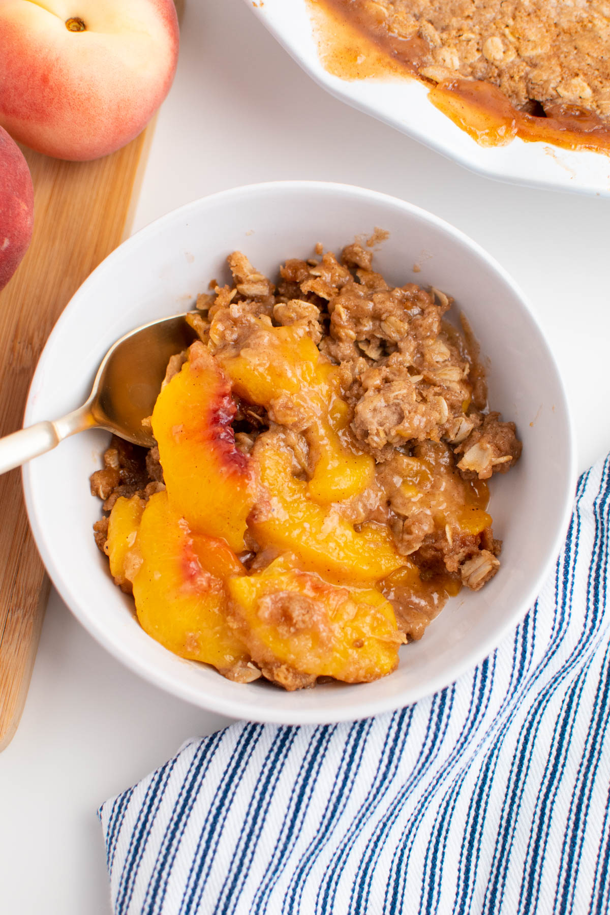 White bowl of peach crisp with spoon and blue striped towel nearby.