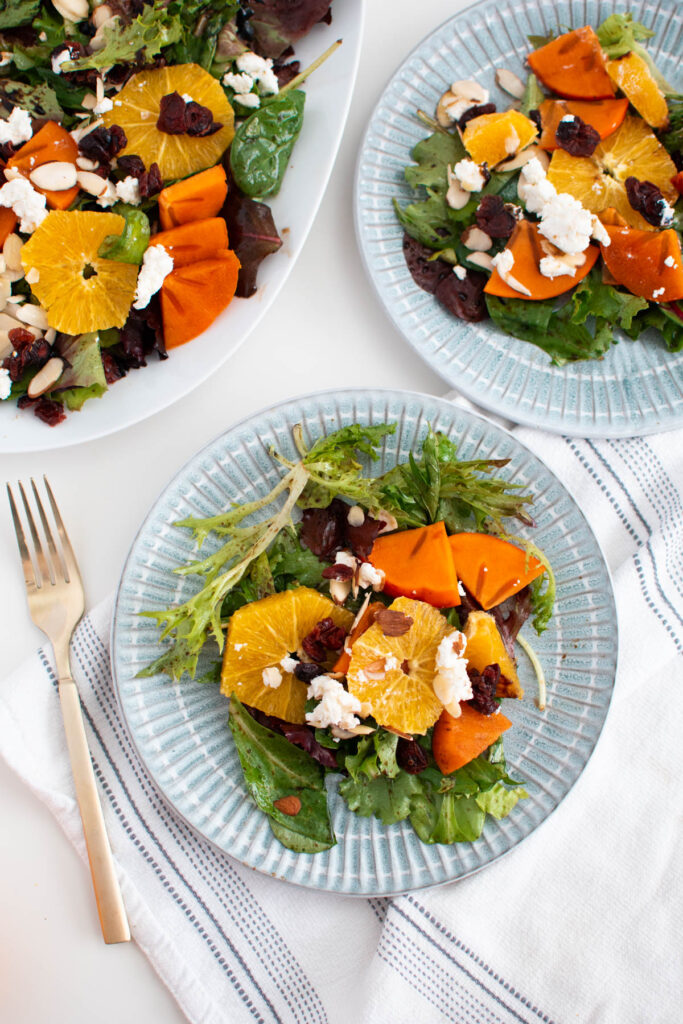 Two blue plates with persimmon goat cheese salad resting on white kitchen towel.