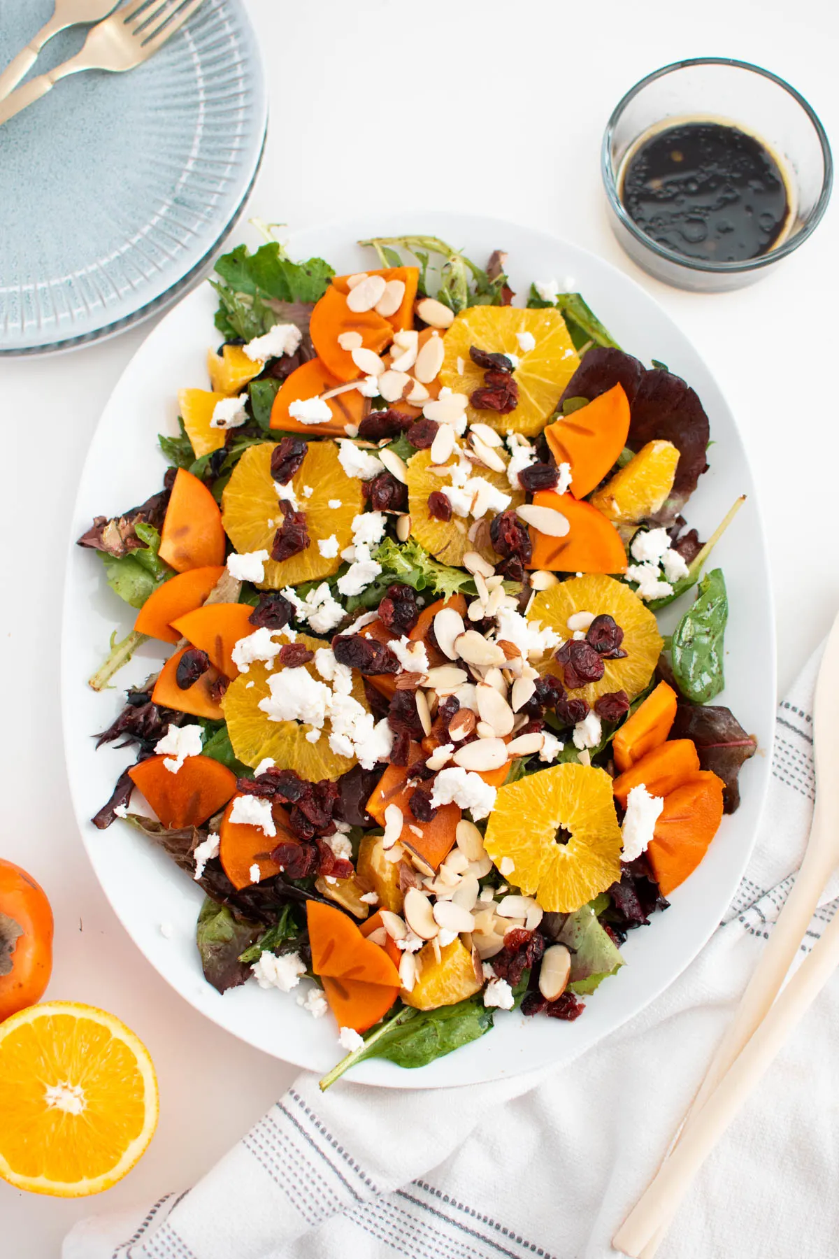 Persimmon salad with oranges, almonds, and cranberries on white platter with plates and fresh ingredients nearby.