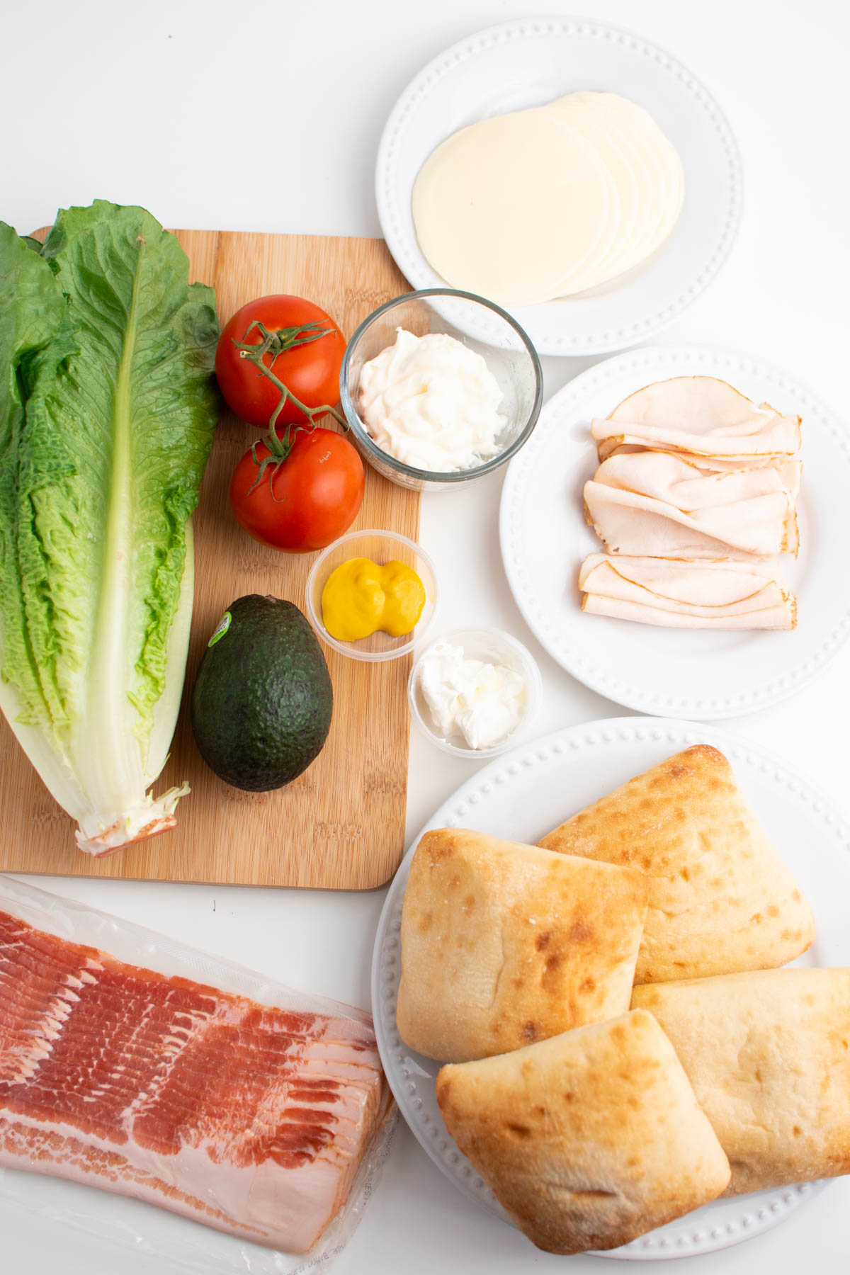 Ingredients on white table including ciabatta rolls, lettuce, deli turkey, and raw bacon.