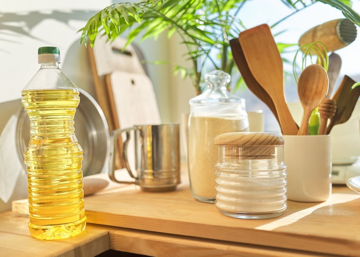 Plastic bottle of yellow oil on wooden countertop next to cooking utensils and glass jars.