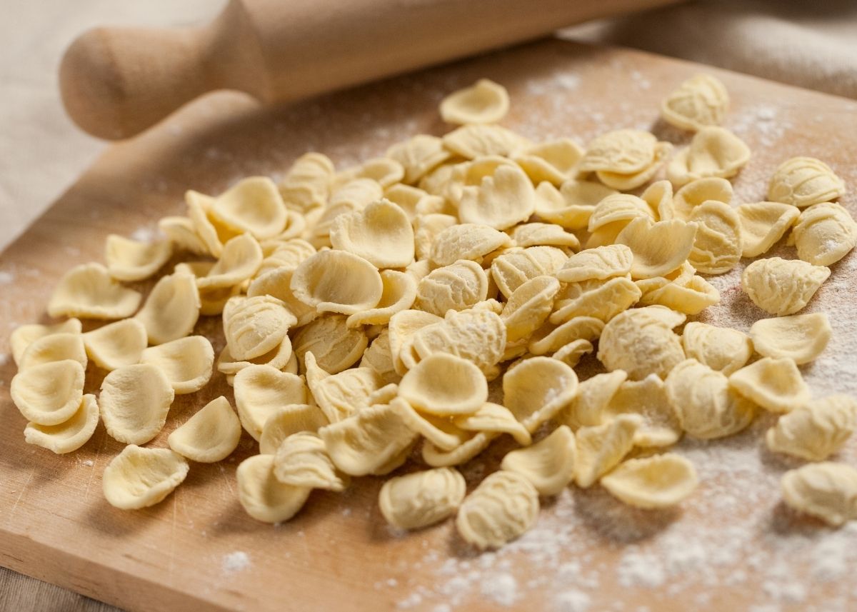 Freshly made, pre-cooked orecchiette pasta next to wooden rolling pin on cutting board.