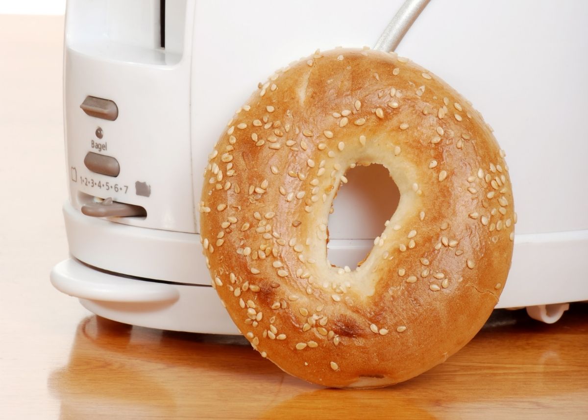 Large fresh bagel propped up on a white and silver toaster on wooden table.