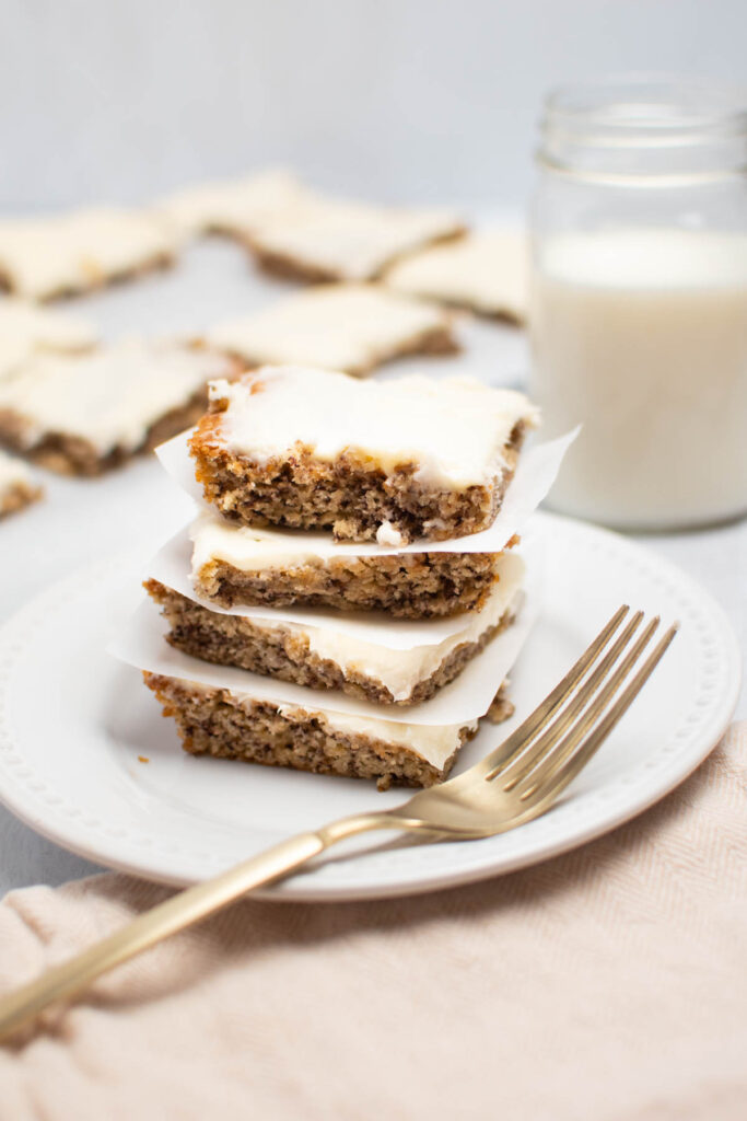 Four banana bars with cream cheese frosting stacked on each other on white plate.