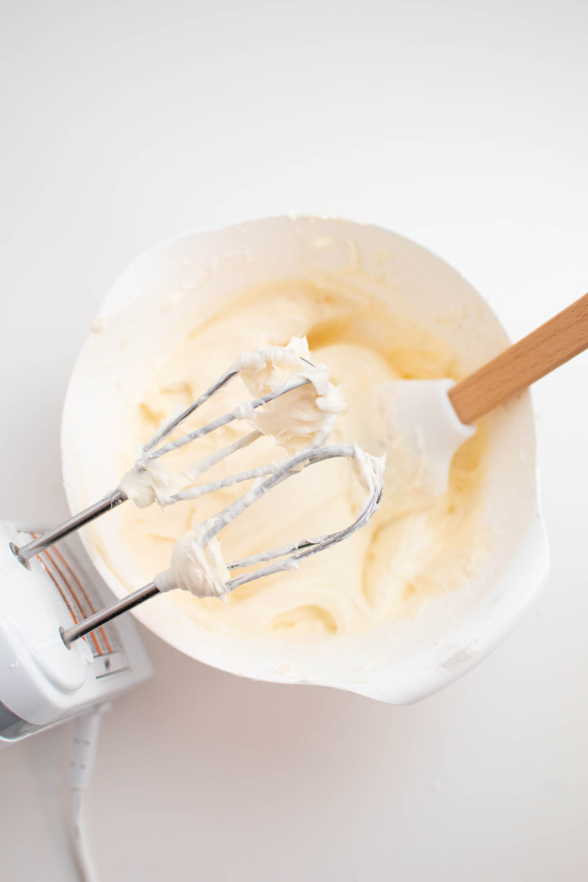 Cream cheese frosting in white bowl with electric beaters above.