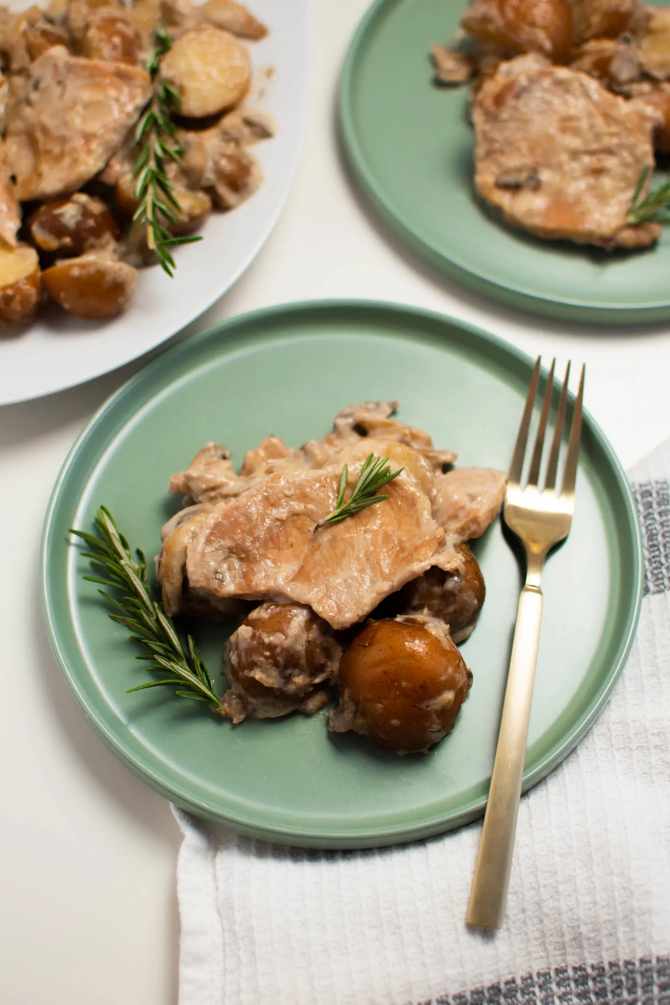 Two green plates with pork chops and red potatoes and sprigs of fresh rosemary on white table.