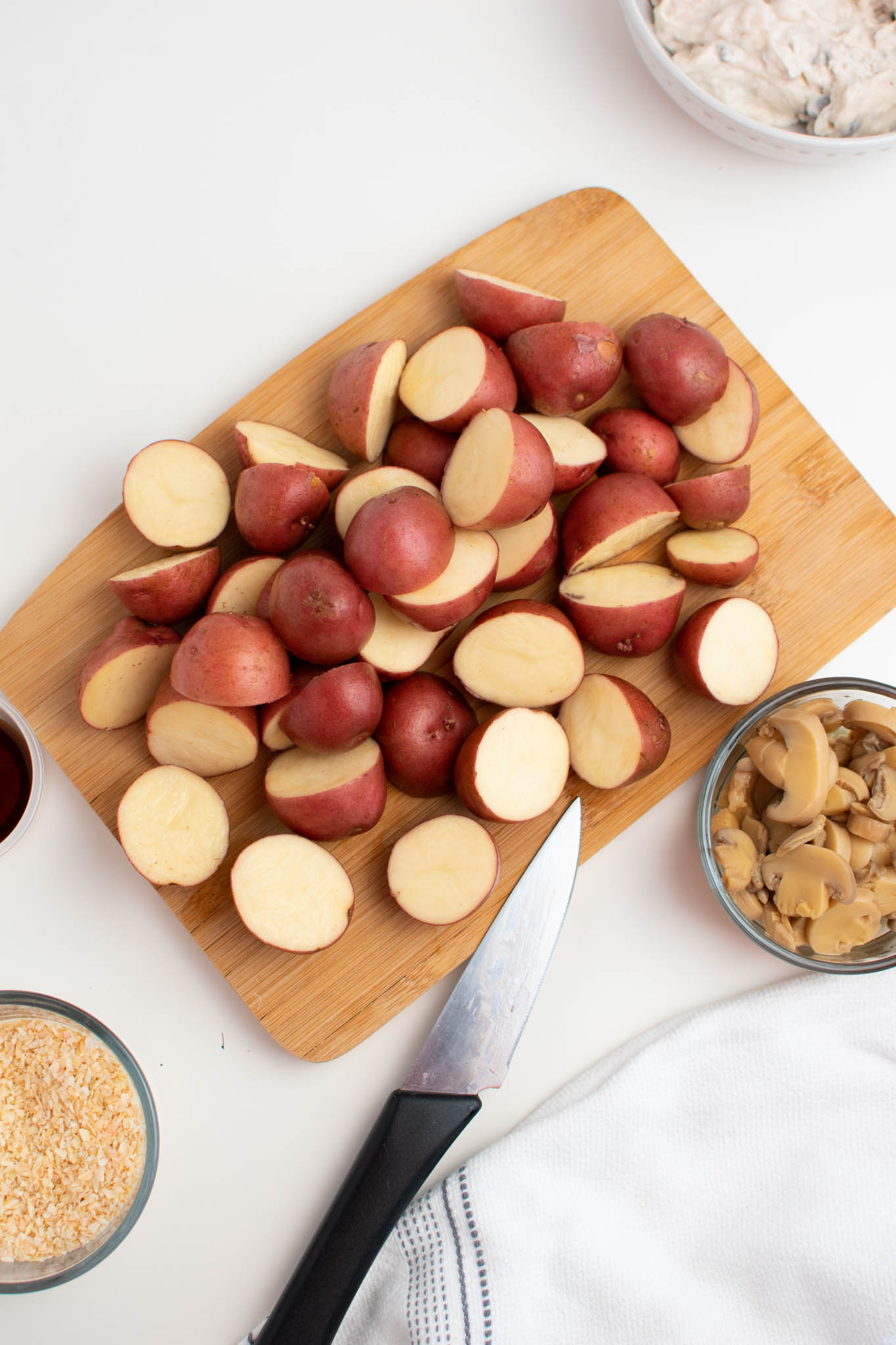 Several cut red potatoes on wood cutting board with bowls of ingredients nearby.