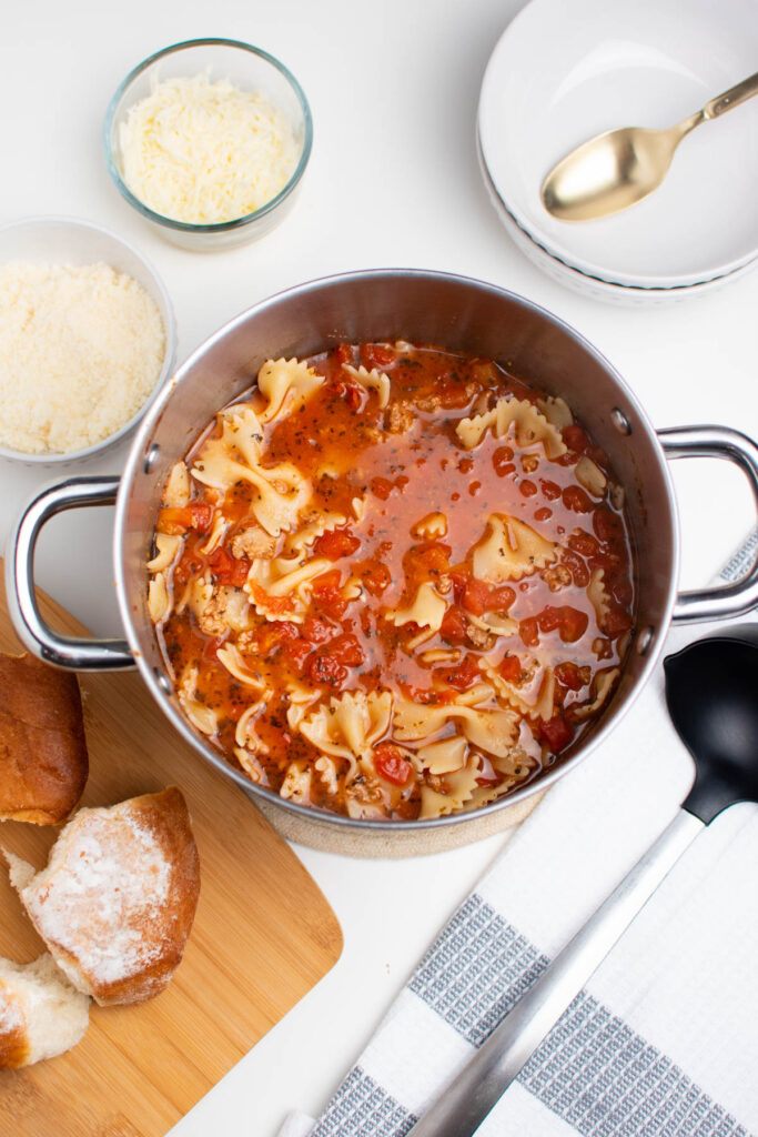 Soup pot full of lasagna soup surrounded by bowls of cheese and rolls on cutting board.
