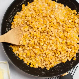 Wooden spoon rests in black frying pan of honey butter skillet corn on white table.