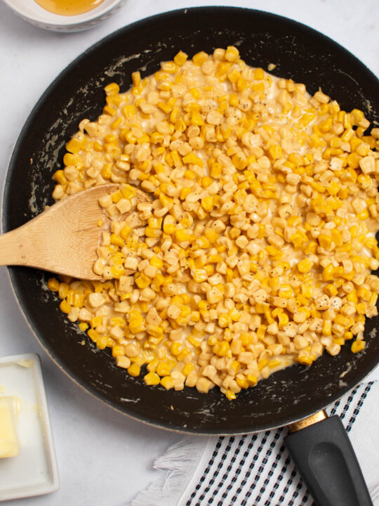 Wooden spoon rests in black frying pan of honey butter skillet corn on white table.