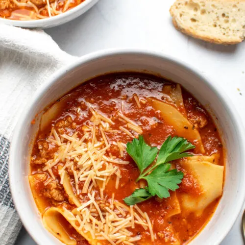 Bowls of lasagna soup on countertop with slices of bread and kitchen towel nearby.