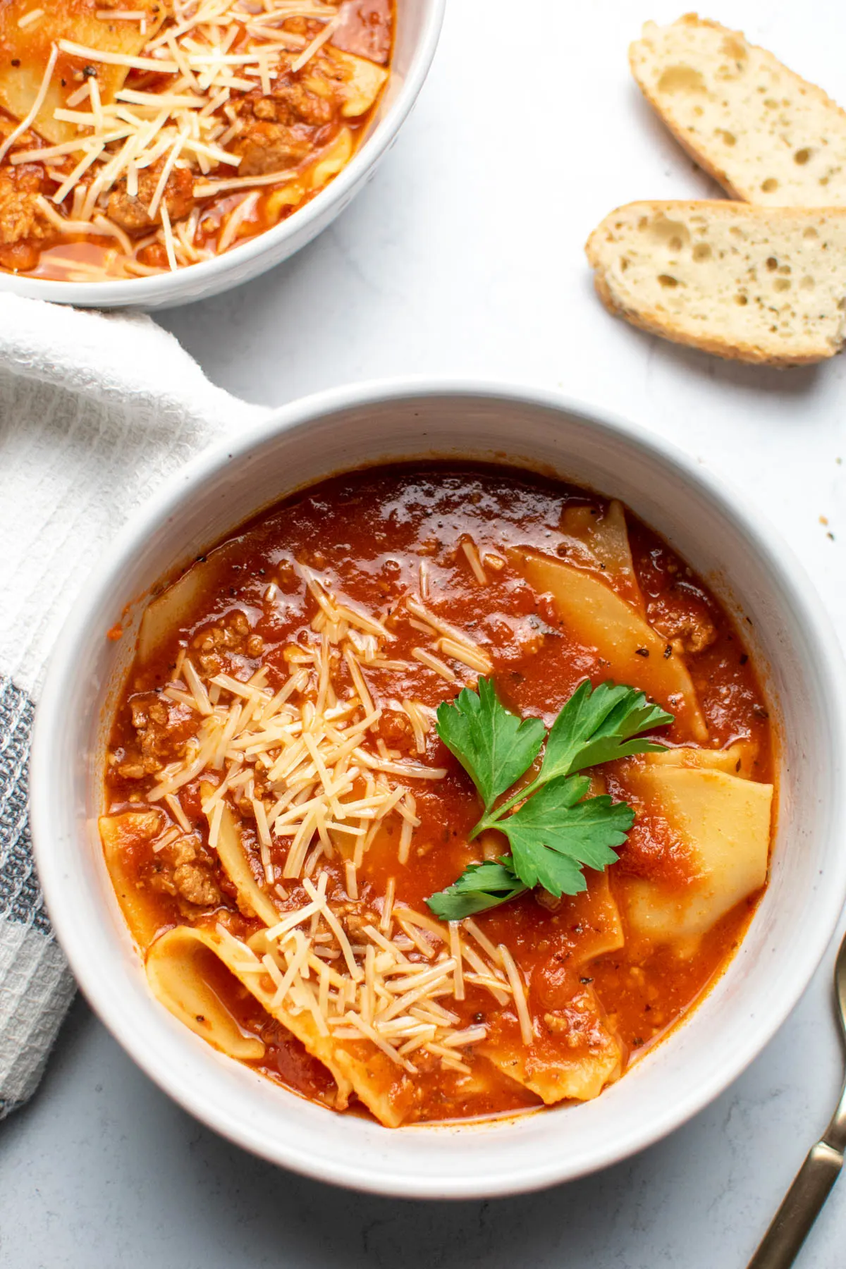 Bowls of lasagna soup on countertop with slices of bread and kitchen towel nearby.