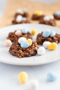 Three birds nest cookies on speckled plate surrounded by pastel coated chocolate eggs.