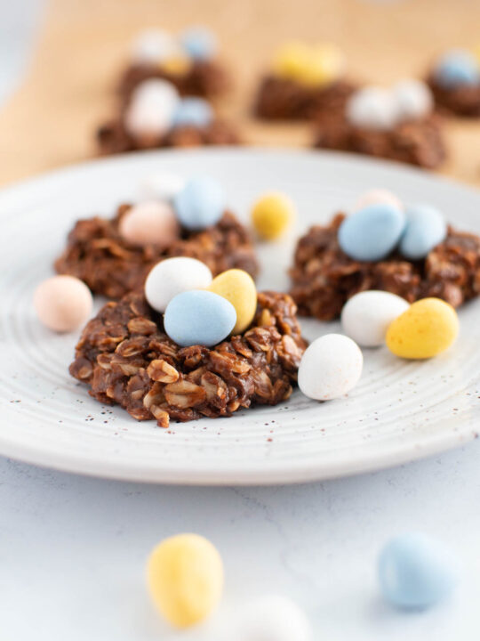 Three birds nest cookies on speckled plate surrounded by pastel coated chocolate eggs.