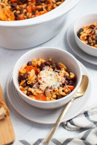 Two white bowls of Crock Pot minestrone soup on plates with gold spoon resting on plate.