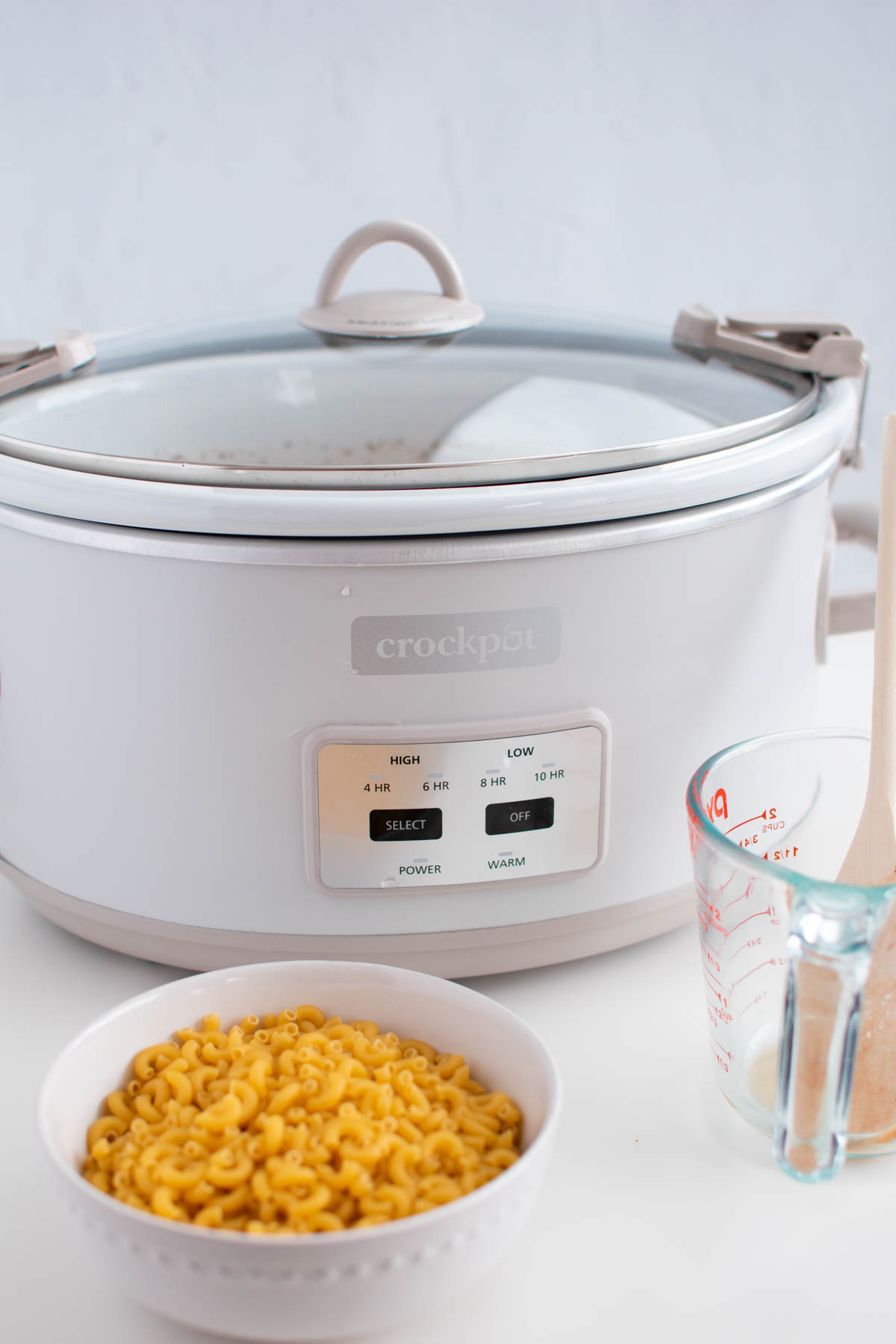 Crock Pot with lid on table with bowl of uncooked macaroni nearby.