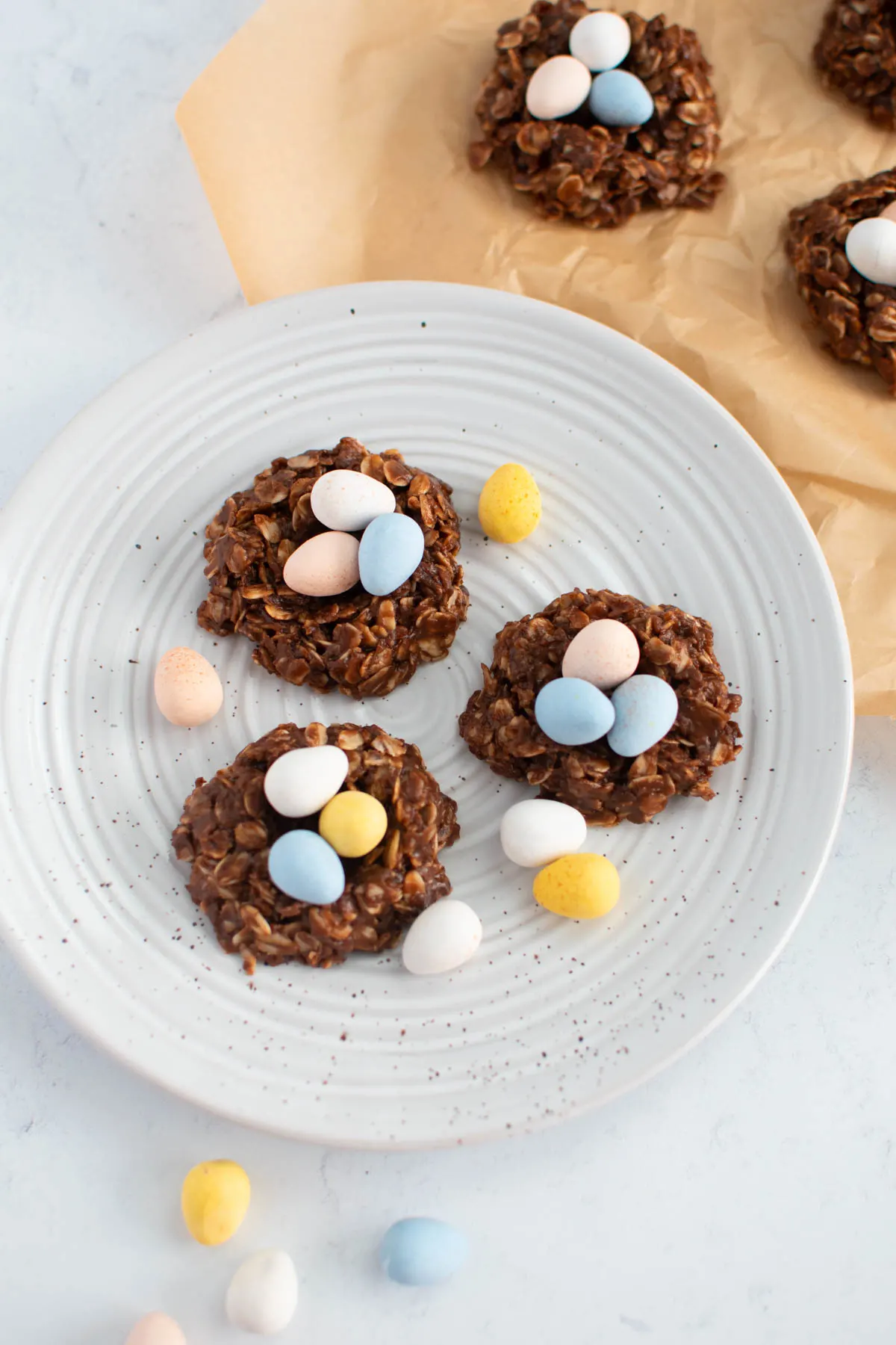 Three Easter nest cookies on speckled plate with loose pastel coated chocolate eggs sprinkled around.