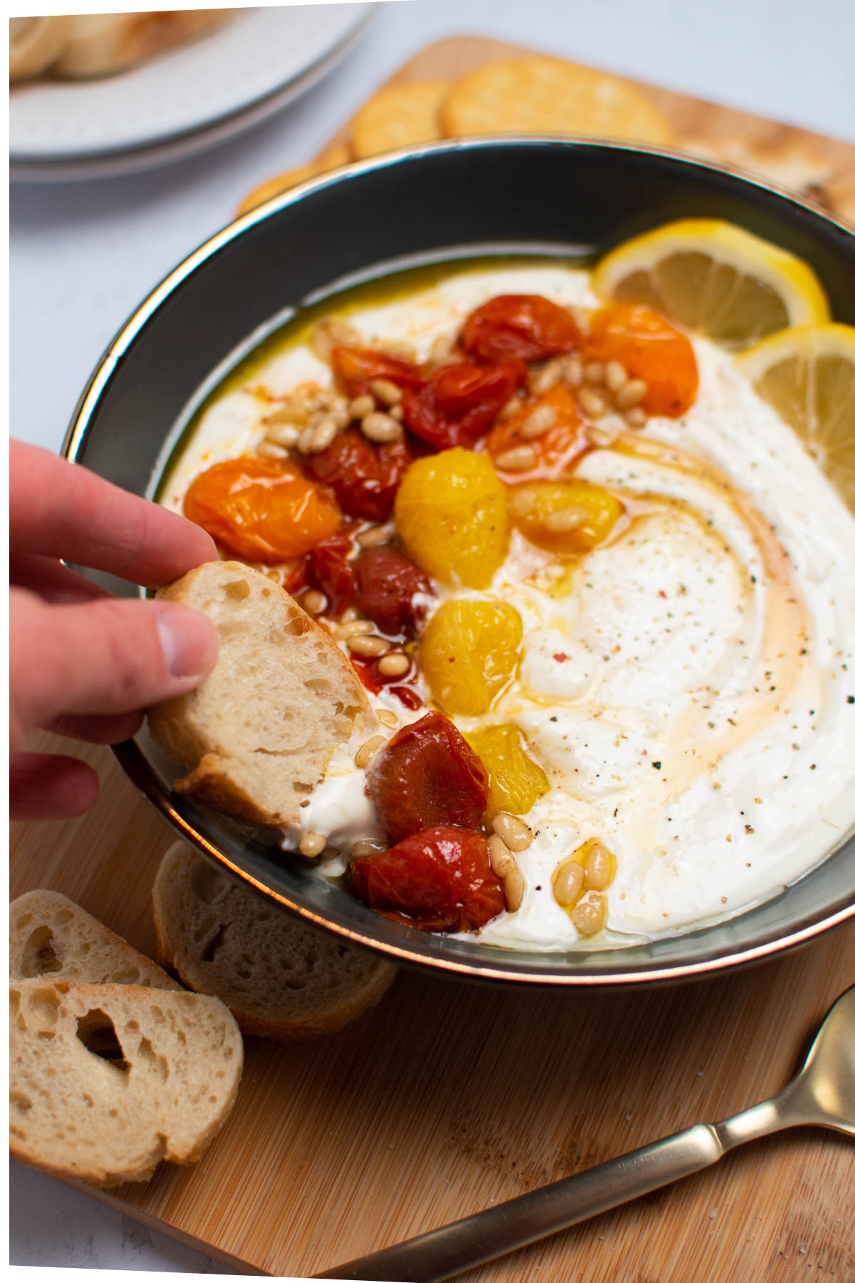 Woman dips slice of baguette in feta cream cheese dip with tomatoes and pine nuts.