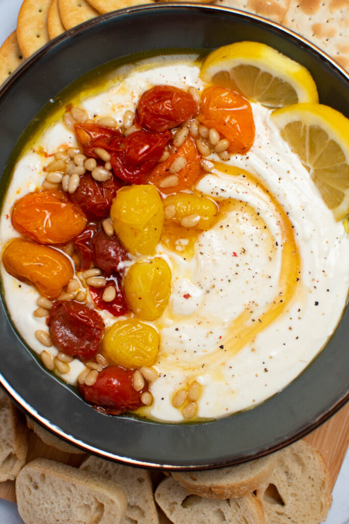 Gray bowl filled with feta dip, roasted tomatoes, pine nuts, honey, and lemon slices.