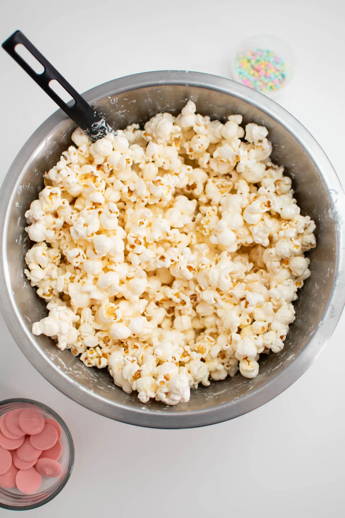 Marshmallow covered popcorn in large mixing bowl with other ingredients nearby.