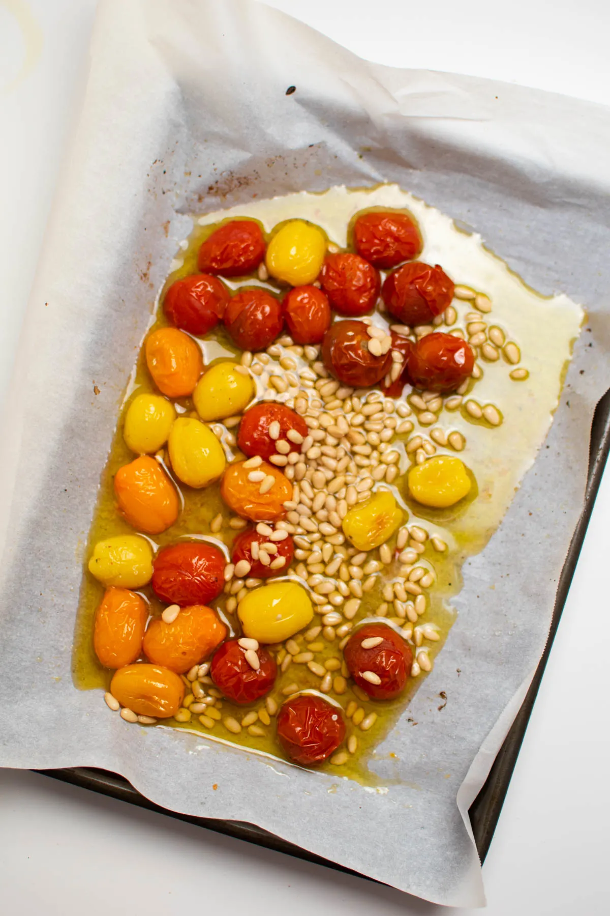 Roasted cherry tomatoes and pine nuts in olive oil on parchment lined baking sheet.