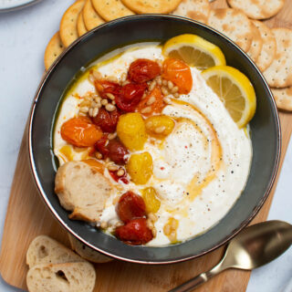 Gray bowl of whipped feta dip with roasted tomatoes surrounded by crackers, slices of bread, and a gold spoon all on wood cutting board.