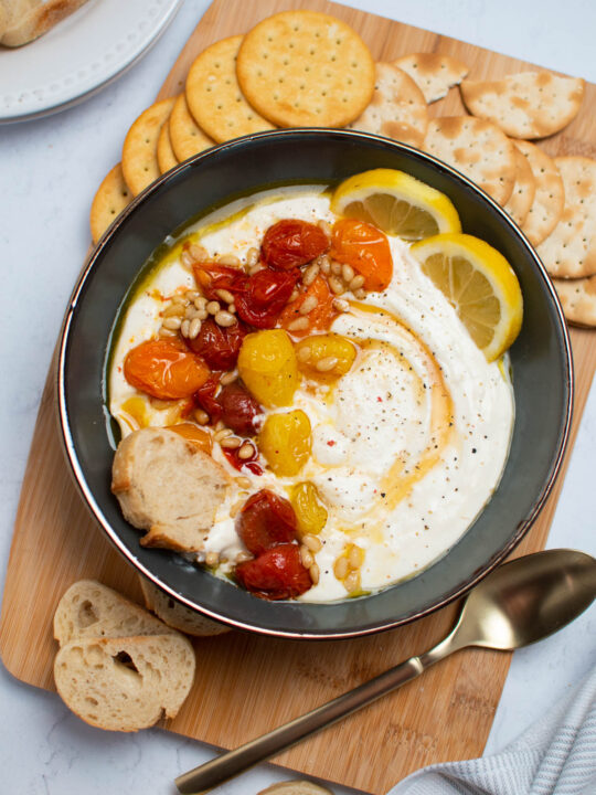 Gray bowl of whipped feta dip with roasted tomatoes surrounded by crackers, slices of bread, and a gold spoon all on wood cutting board.
