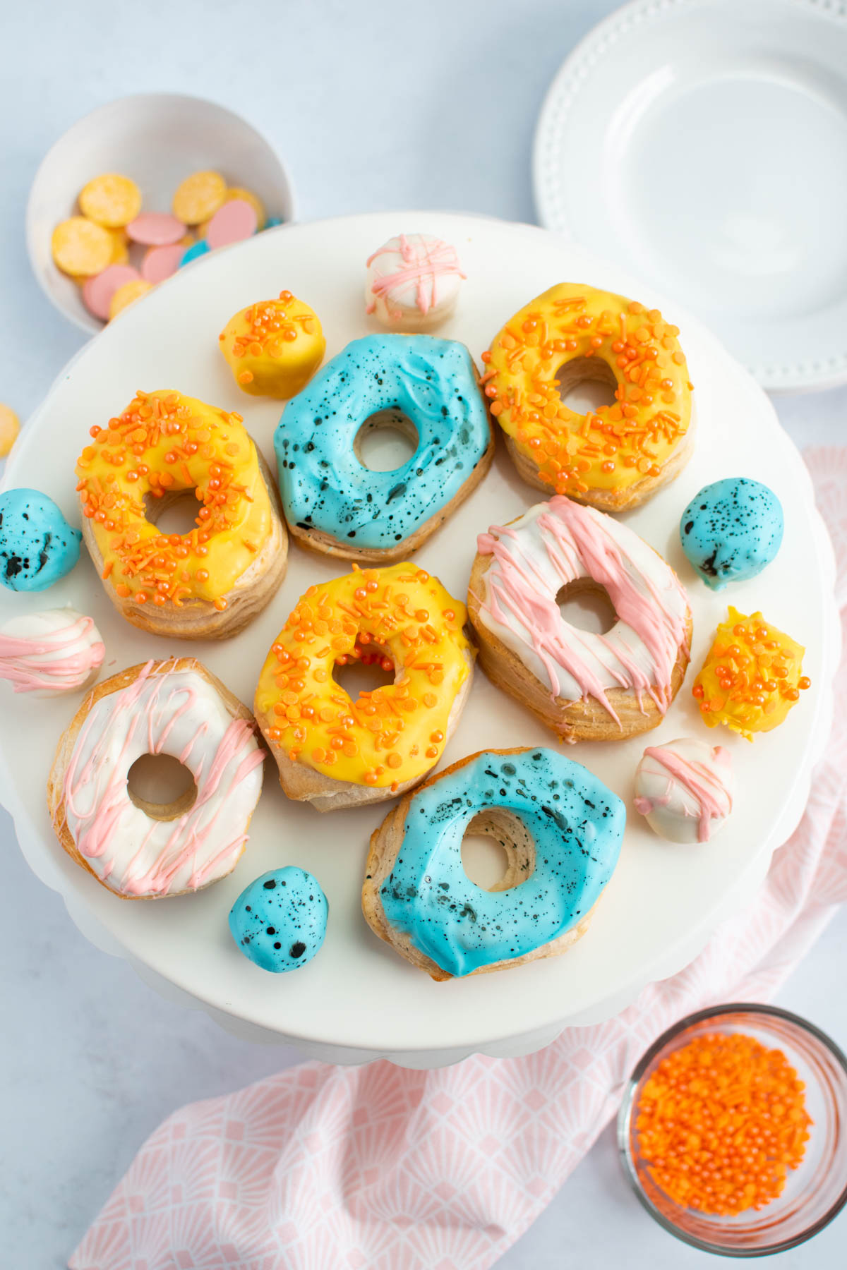 Easter decorated air fryer donuts on cake stand with plates and bowls of sprinkles nearby.