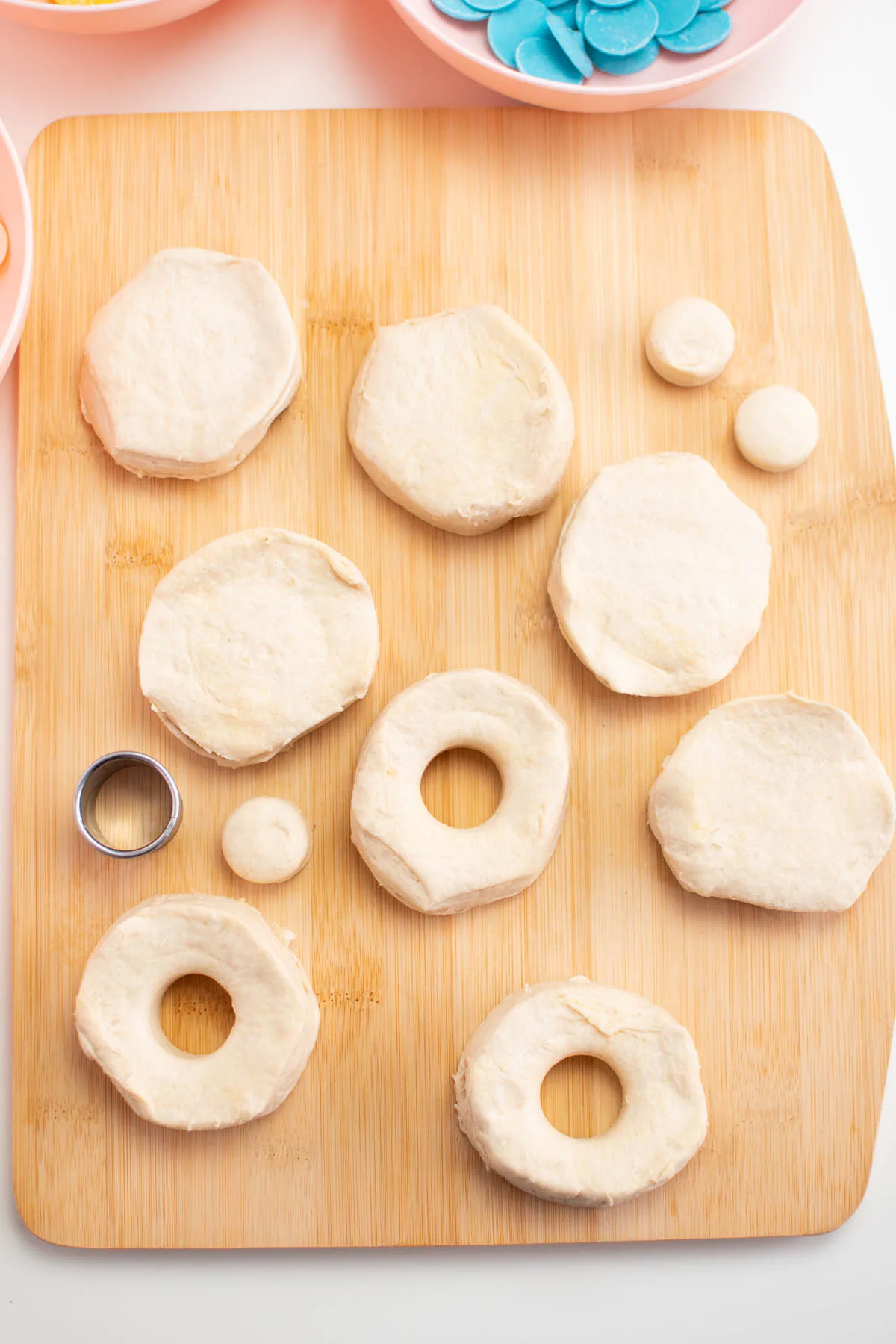 Raw biscuit dough circles with hole cut of center on wood cutting board.