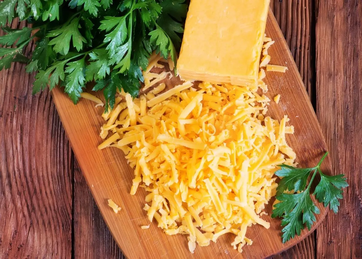Cheddar cheese block and shreds with parsley on wooden cutting board.