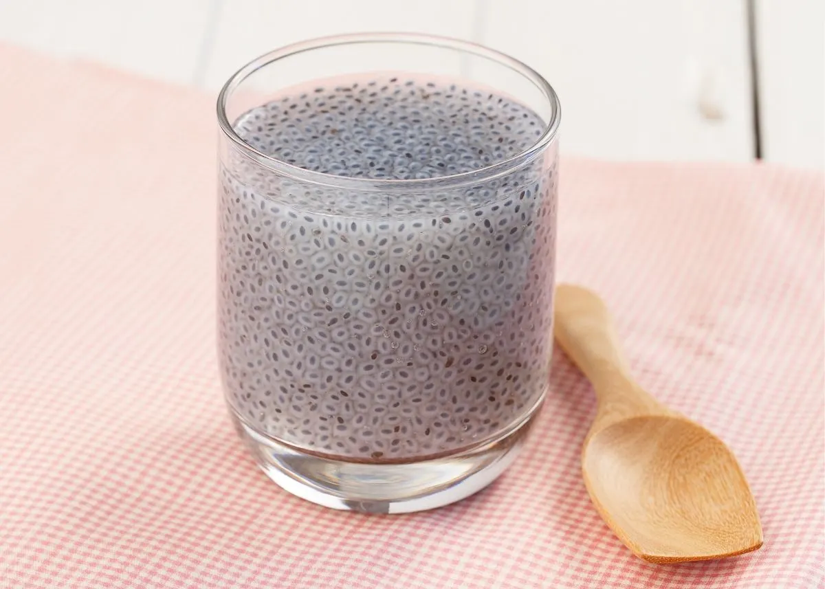 Large glass of water with chia seeds next to wooden spoon on checkered tablecloth.