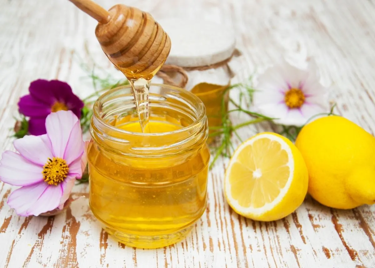 Honey spills from stirrer into jar, surrounded by flowers and lemons.