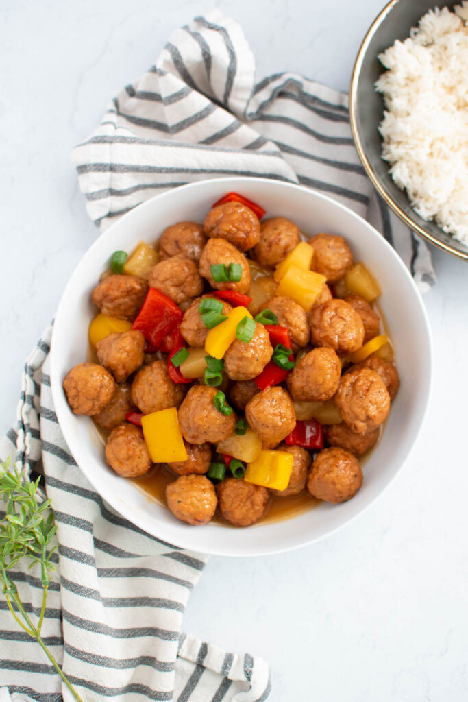 White bowl of Instant Pot sweet and sour meatballs with pineapple and bell peppers with bowl of cooked rice nearby.
