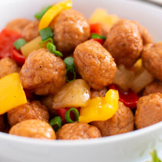 White bowl full of sweet and sour meatballs, bell peppers, pineapple, and green onions.
